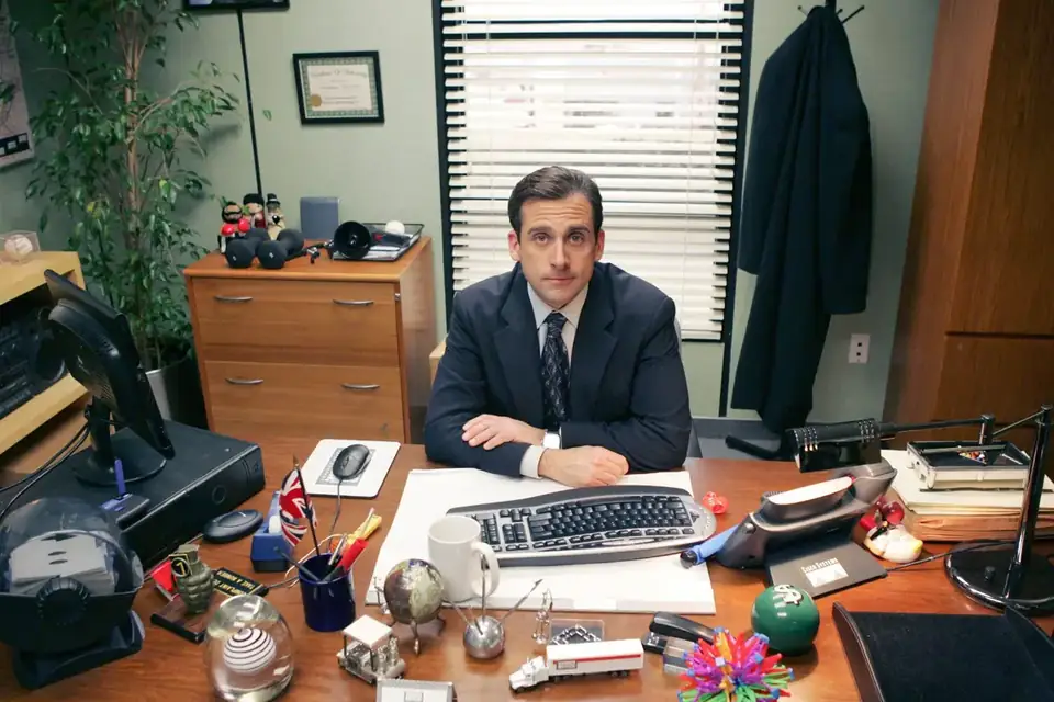 Steve Carell as Michael Scott sitting behind his desk in The Office